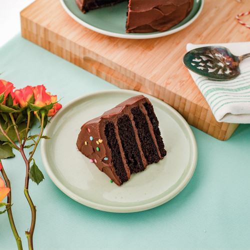 Image 2: Chocolate cake slice shown on a table so you can visually see the 4 layers of moist chocolate cake with the chocolate buttercream. View 2